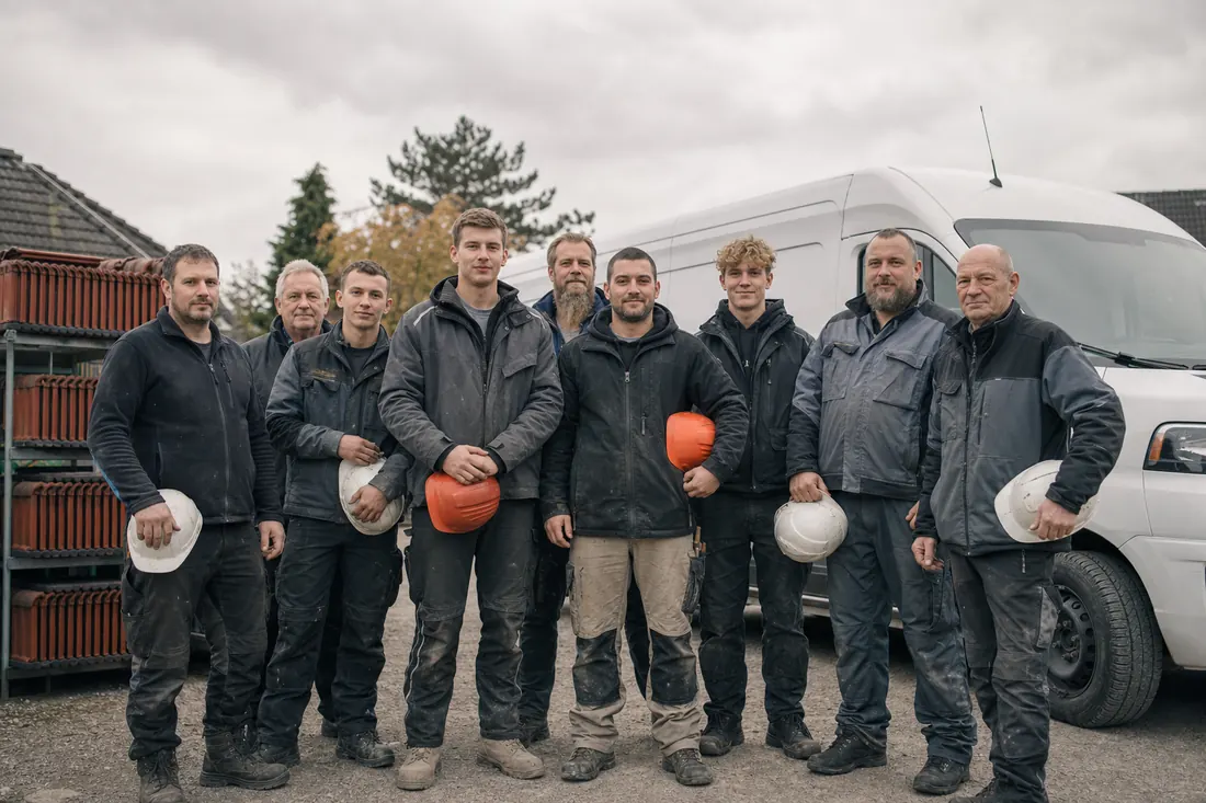 Neun Dachdecker in Arbeitskleidung stehen vor einem Transporter und einem Stapel Tonziegel